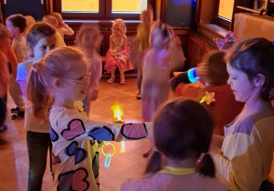 dzieci tańczą i bawią przy zasłoniętych oknach, aby widać było neonowe bransoletki i inne ozdoby fluorescencyjne