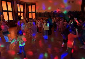 dzieci tańczą i bawią przy zasłoniętych oknach, aby widać było neonowe bransoletki i inne ozdoby fluorescencyjne