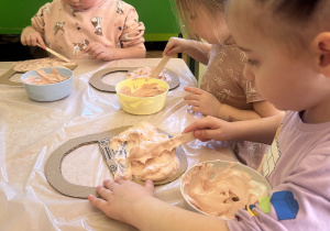 Na zdjęciu dzieci przy stoliku nakładają na tekturowe koszyczki masę plastyczną z pianki do golenia i farby.