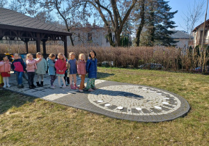 Grupa dzieci stoi przed altaną, przed dziećmi na kamiennym zegarze leżą poukładane kropelkowe memory