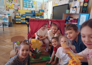 dzieci siedzą przed przygotowanym przez siebie teatrzyku czyli stojaka z dwiema zasłonkami imitującymi kurtynę