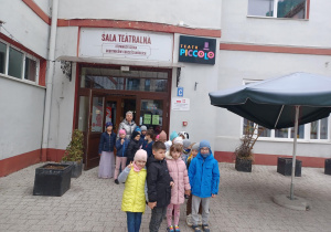 Grupa dzieci stoi na dziedzińcu przed wejściem do teatru Piccolo. nad wejściem widać duży napisy: Sala Teatralna i Teatr Piccolo