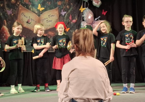 dzieci z instrumentami na scenie, przed nimi nauczycielka , która dyryguje