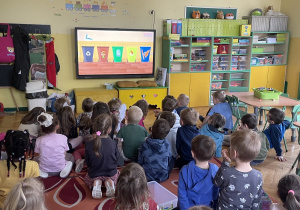Dzieci z grupy Pszczółek oraz Misiów siedzące tyłem na dywanie, oglądające film edukacyjny dotyczący segregacji odpadów