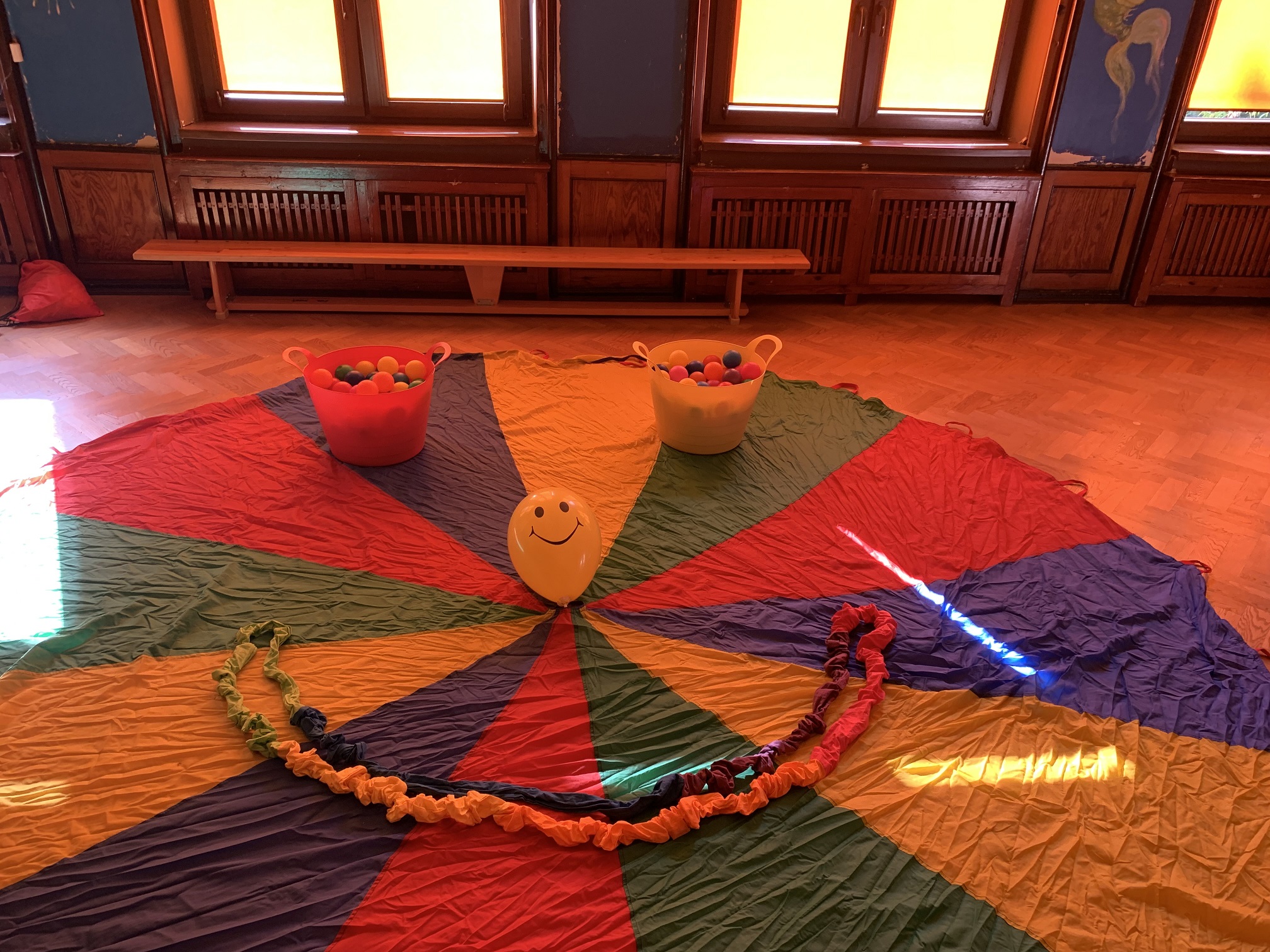 na zdjęciu - rozłożona na podłodze chusta animacyjna , na niej 2 pojemniki z kolorowymi piłkami, balonik i Siguma ułożone w uśmiechnietą minkę.