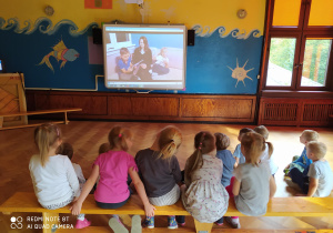 Dzieci siedząc na ławce słuchają bajki czytanej przez Brata Boryska i jego mamę.