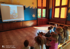 Przedszkolaki siedząc na ławce słuchają bajki czytanej przez Olka i jego mamę.