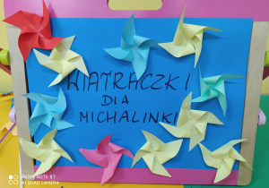 Zdjęcie przedstawia kolorowe papierowe wiatraczki przyklejone na niebieskim kartonie z napisem "wiatraczki dla Michalinki"