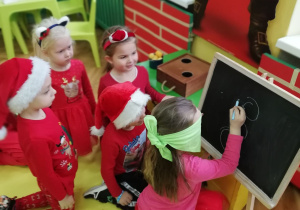 Zdjęcie przedstawia czworo dzieci z grupy "Pszczółek" siedzących na podłodze, jedna z dziewczynek z zawiązaną chustą na oczach w kolorze zielonym rysuje na tablicy.