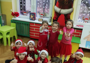 Zdjęcie przedstawia przedszkolaki w czerwonych strojach oraz "Mikołajkowych" czapkach siedzących na podłodze przed tablicą z rysunkiem Mikołaja podnoszących kciuki w górę.