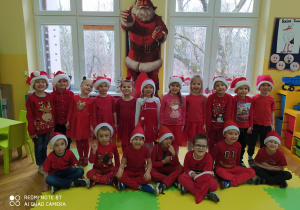Zdjęcie przedstawia dzieci z grupy "Pszczółek" w czerwonych strojach oraz "Mikołajkowych" czapkach