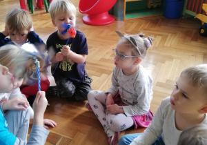 Na zdjęciu dzieci siedzą na podłodze, chłopiec dmucha w wiatraczek.