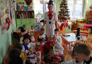 Na zdjęciu znajdują się dzieci z nauczycielką przebrani w stroje bałwanków siedzą przy stole zastawionym słodyczami i owocami