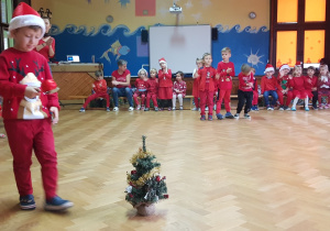 na zdjęciu chłopiec, który niesie bombkę choinkową na łyżce, w tle dzieci, które siedzą na ławeczce i dopingują chłopcu, dzieci w strojach z dominującym kolorem czerwonym