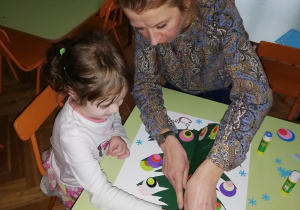 na zdjęciu dziewczynka z nauczycielka przykleja pawie oczko na papierowej choince