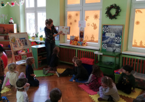 na zdjęciu - widok klasy przedszkolnej, dzieci siedzące na podłodze patrzą na nauczycielkę, która pokazuje książeczkę o Kubusiu Puchatku