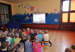widok sali, na której siedzą dzieci na ławeczkach i oglądają bajkę na tablicy multimedialnej