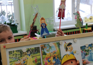 Na zdjęciu dzieci bawią się w teatr - na zdjęciu widać stojąca niską tablicę, na której przyczepione są różne obrazki z bajek, nad nią widać rączki dzieci trzymające papierowe kukiełki do bajki "O dzielnym Szewczyku".