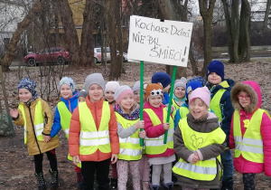 Na zdjęciu grupa dzieci stojących w parku, w tle domy jednorodzinne. Dwoje dzieci z przodu grupy trzyma transparent z napisem "Kochasz dzieci, nie pal śmieci"