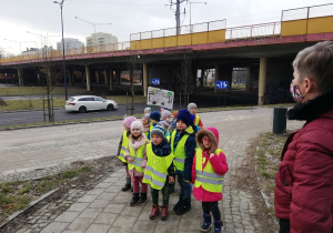 Na zdjęciu grupa dzieci stojących na chodniku, w tle wielopasmowa ulica i jadące samochody. Dwoje dzieci na czele grupy trzyma transparent z napisem "Stop smogowi".