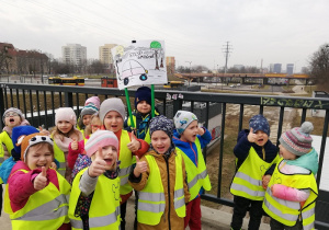 Na zdjęciu grupa dzieci stojących na kładce łączącej park z ulicą Tuwima. W tle widać zajezdnię autobusowa oraz wielopasmową ulicę i jadące samochody. Dwoje dzieci na czele grupy trzyma transparent z napisem "Stop smogowi", pozostałe dzieci pokazują kciuk znaczący "jest ok"