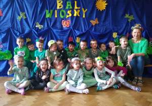 Zbiorowe zdjęcie grupy dzieci wraz z nauczycielką na tle dekoracji z napisem bal wiosny