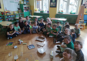 Grupa przedszkolaków siedzi na podłodze, przed dziećmi na podłodze znajdują się surowce wtórne : papier, plastik, szkło.