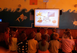 Dzieci z grupy VI i V siedzą na ławeczkach w sali gimnastycznej i oglądają film edukacyjny o Unii Europejskiej.