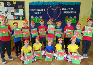 Odświętnie, kolorowo ubrane starszaki z grupy VI na tle dekoracji związanej z Dniem Mamy i Taty prezentują wykonane w przedszkolu laurki dla swoich Rodziców.