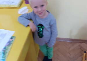 Na zdjęciu uśmiechnięty przedszkolak stojący przy biurku nauczycielki.