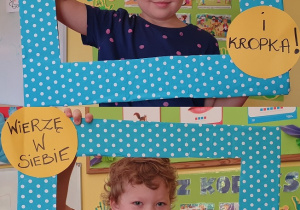 Na zdjęciu dwoje dzieci pozujących w niebieskiej ramce w kropki z napisem :Wierzę w siebie.. i kropka!"