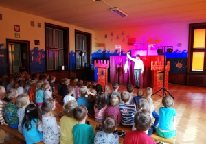 dzieci oglądają występ aktorów, w tle scenografia do przedstawienia i aktor pokazujący marionetkę