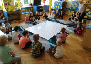 dzieci siedzą na dywanie, przed nimi mata do kodowania z zaznaczonymi kropkami