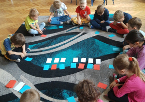 Dzieci przedszkole siedzą na dywanie w klasie w siadzie skrzyżnym. Uczestniczą w zajęciach w zakresie analizy i syntezy wyrazów z wykorzystaniem nakrywek w trzech kolorach; białym, czerwonym i niebieskich. Na zdjęciu widoczne skupienie i chęć współpracy podczas wykonywania zadania.