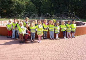 zdjęcie przedstawia dzieci wraz z nauczycielką stojące dookoła parkowej fontanny