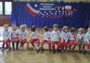 Dzieci z grupy pierwszej siedzące na ławce w oczekiwaniu na rozpoczęcie uroczystości na tle dekoracji z okazji 11 listopada