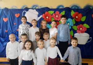 Grupa dzieci przedszkolnych ubranych w stroje galowe, pozuje do zdjęcia na tle dekoracji z kolorowych kwiatów przygotowanej z okazji Dnia Babci i Dziadka.