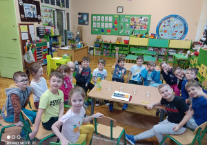 Na zdjęciu grupa dzieci siedzi na krzesełkach obserwują materiały potrzebne do przeprowadzenia eksperymentu wody z olejem i kolorowymi kostkami lodu.