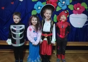 Grupka uśmiechniętych dzieci przedszkolnych ubranych w kolorowe stroje karnawałowe przedstawiające różne postaci z bajek, pozują do zdjęcia na tle dekoracji przedstawiającej dzbanek z filiżanką do kawy i kolorowe piękne kwiaty.