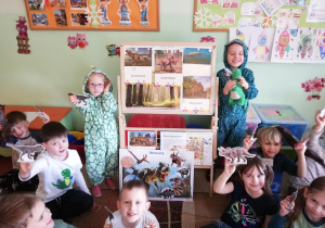 grupa dzieci siedząca na dywanie i pokazująca wykonane prze siebie tekturowe dinozaury. dzieci sa ubrane w odzież z nadrukami przedstawiającymi różne dinozaury. Dwoje dzieci jest przebrane za dinozaury