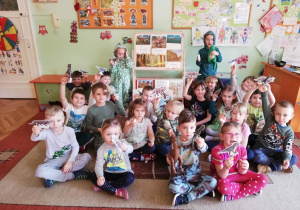 grupa dzieci siedząca na dywanie i pokazująca wykonane prze siebie tekturowe dinozaury. dzieci sa ubrane w odzież z nadrukami przedstawiającymi różne dinozaury. Dwoje dzieci jest przebrane za dinozaury