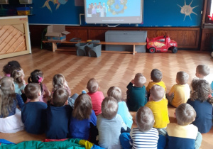 Na zdjęciu dzieci siedzące na podłodze na sali gimnastycznej oglądające film edukacyjny.