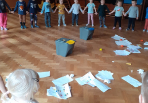 Na zdjęciu dzieci trzymające się za ręce stojące w kole wokół porozrzucanych śmieci, które będą segregować.