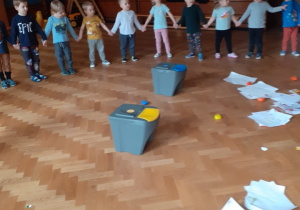 Na zdjęciu dzieci trzymające się za ręce stojące w kole wokół porozrzucanych śmieci, które będą segregować.