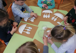 zieci przy stolikach przygotowują prace plastyczną " Ptaki-pisklaki w gniazdach" z wykorzystaniem materiałów nietypowych (m.in. plastikowe łyżeczki, talerzyki papierowe, sianko)