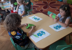 na zdjęciu dzieci siedzące przy stolikach wykonujące pracę plastyczna – wycinanie nożyczkami, wyklejanie plasteliną sylwety kwiatów.