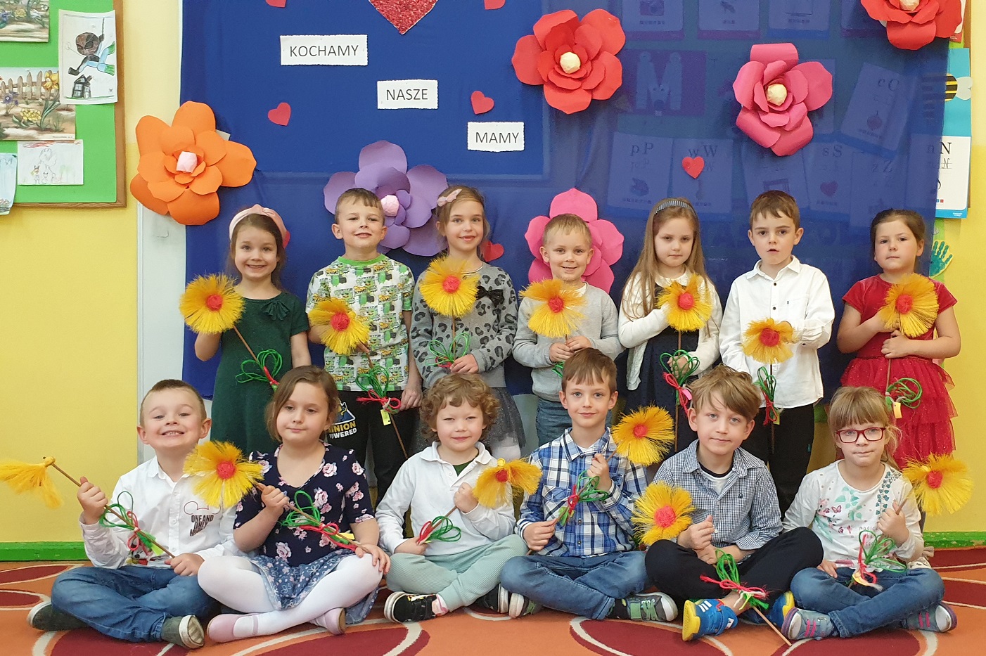 Na zdjęciu dzieci z grupy Misiów pozują z własnoręcznie wykonanymi ze sznurka kwiatami na niebieskim tle z napisem "kochamy nasze mamy"