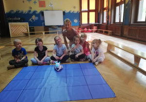 dzieci wraz nauczycielką siedzą przed matą do kodowania, na której stoi mały robocik