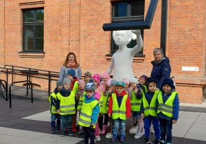 Zdjęcie grupowe dzieci i opiekunów stojących przy rzeźbie białej dziewczynki, na tle budynku z czerwonej cegły.