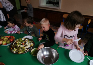 Dzieci na jadalni przy wspólnym stole kroją plastikowymi nożykami na małych talerzykach owoce na sałatkę